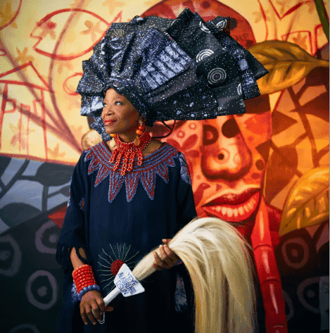 Woman in traditional African attire with elaborate headpiece and art mural background.
