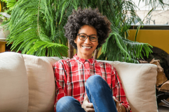 African woman with curly hair and glasses smiling on a sofa.