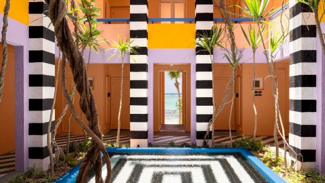 Colorful tropical courtyard featuring bold striped columns and lush plants, with a view of the ocean.
