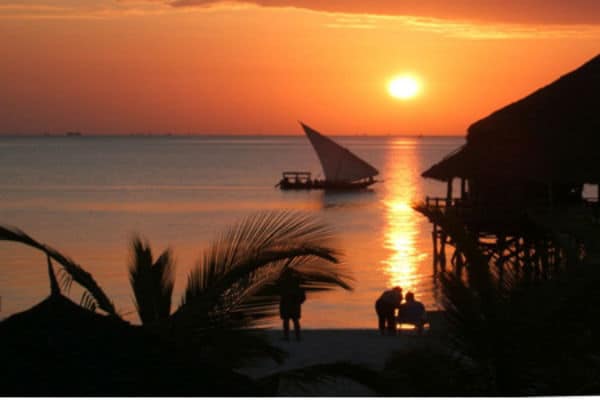 Sunset over calm waters with a traditional sailboat and silhouetted palm trees in Africa.