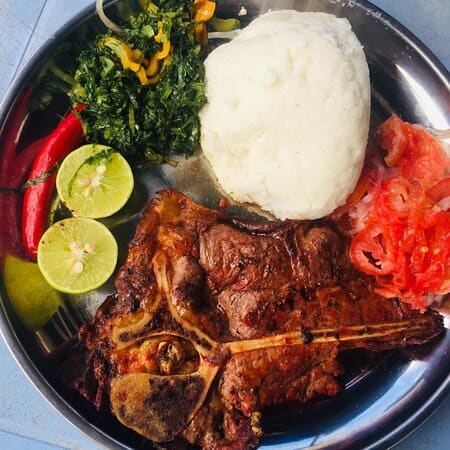 A plate of Nigerian cuisine featuring grilled meat, mashed potatoes, fresh tomatoes, and greens for.