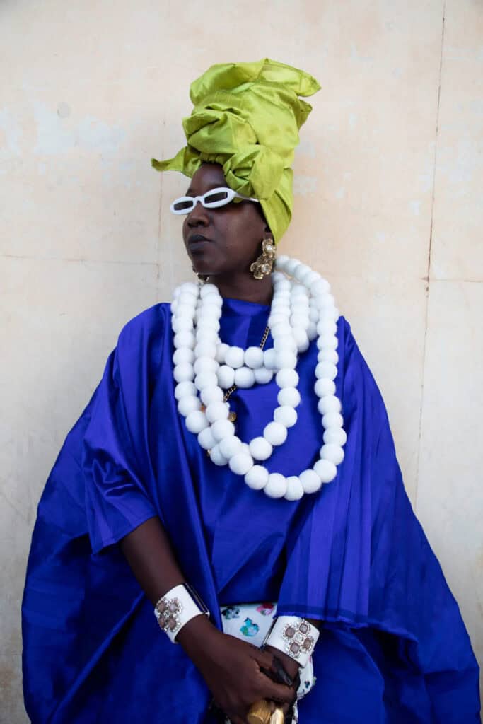 African woman in colorful traditional attire with headwrap, jewelry, and sunglasses.