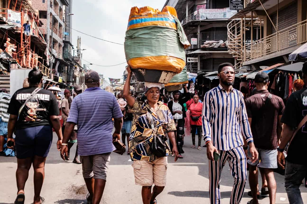 A woman carrying a large load on her head walks through a busy African marketplace.