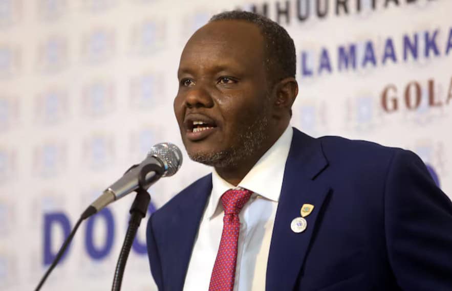 African leader speaking at a conference, wearing a navy suit and red tie.