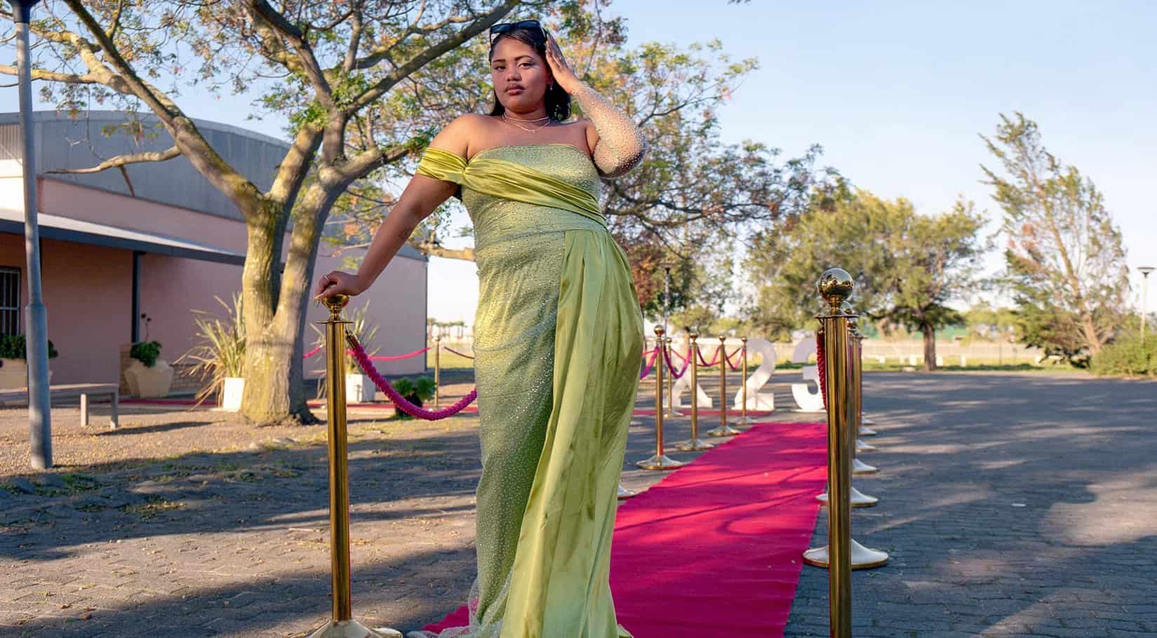 Woman in green dress walking on red carpet outdoors, surrounded by trees and clear sky.